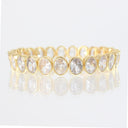 A gold-plated stretch bracelet featuring a row of oval-cut clear crystals in bezel settings, displayed on a reflective white surface.