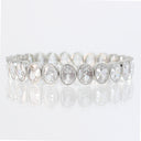A silver-toned tennis bracelet featuring a row of oval-cut clear gemstones in a prong setting, displayed on a reflective white surface against a white background.
