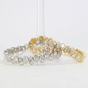 Two tennis-style bracelets with teardrop-shaped clear stones, one in silver-tone metal and one in yellow gold-tone metal, displayed on a clear stand against a white background.