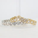 Two tennis-style bracelets stacked together on a clear display stand, one in yellow gold and one in silver, both featuring round clear gemstones in bezel settings.