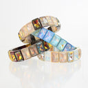 Three iridescent square-rimmed bangles stacked together on a reflective white surface. The bangles feature shifting colors including gold, pink, blue, and green, set in a dark metallic frame.