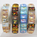 Four rigid bangle bracelets with rectangular faceted rhinestones, displayed side by side on a white stand. The bracelets feature different color combinations and finishes: clear/iridescent, yellow-gold/amber, light blue/teal, and peach/orange iridescent.